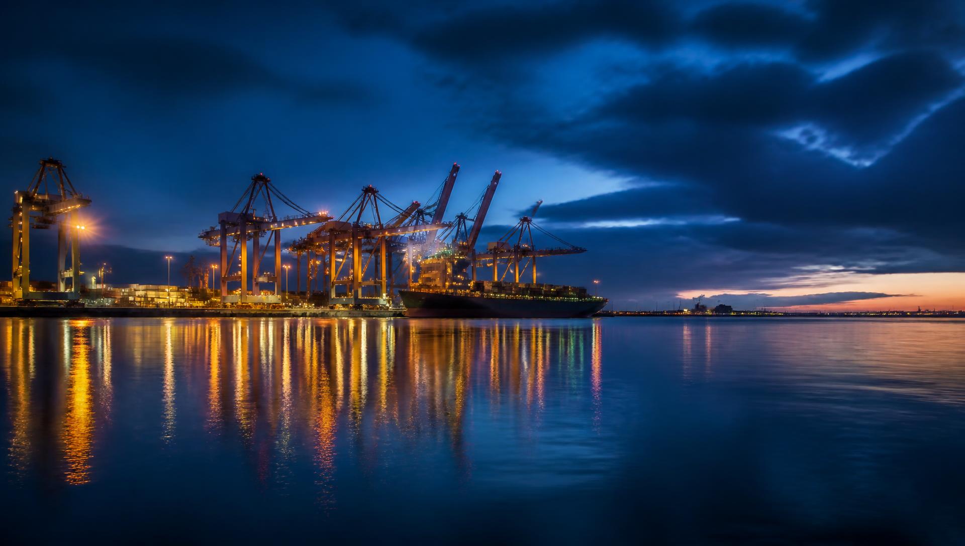 Illuminated port cranes and a cargo ship reflected on calm water at blue hour